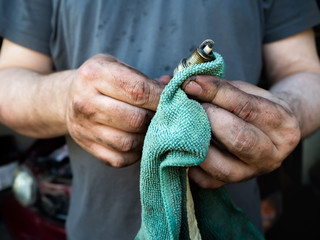 a man in dirty clothes and with grubby hands, holding a rag and a spark plug, wiping the car parts