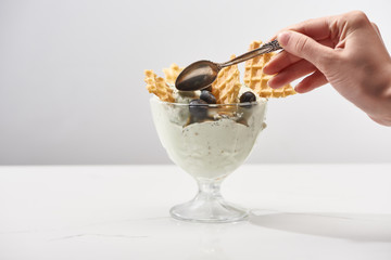 cropped view of woman holding spoon near bowl with pistachio ice cream with waffles and blueberries isolated on grey