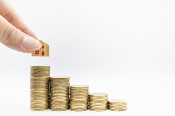 Home loan, business, money and saving concept. Close up of hand holding wooden house toy and putting down on top of stack of gold coins on white background and copy space.