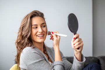 Woman with brush and mirror on hands put make up on face while sitting on sofa
