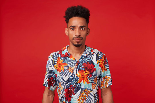 Young wondered African American man, wears in Hawaiian shirt, looks at the camera with surprised expression and wide opened eyes, stands over red background.