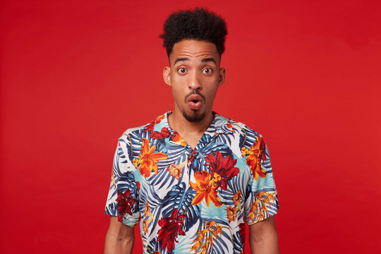 Young surprised African American man, wears in Hawaiian shirt, looks at the camera with amazed expression and wide opened eyes, stands over red background.