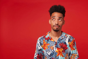 Young pensive African American guy wears in Hawaiian shirt, looking up, thinking and imagine, stands over red background with copy space at the left side.