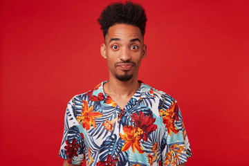 Young confused African American guy wears in Hawaiian shirt, looking at the camera with wondered and misunderstanding, stands over red background with crossed arms.