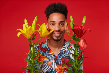 Young African American guy, wears in Hawaiian shirt, looks at the camera with happy expression, stands over red background with yellow and red flowers and smiles.