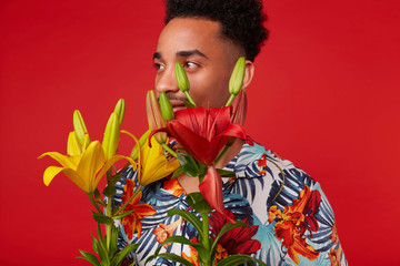 Close up of young African American guy, wears in Hawaiian shirt, looks away, stands over red background, yellow and red flowers covered his face.