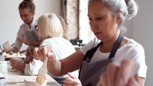 Medium shot of middle-aged mixed-race woman painting clay mug with brush while her teammates sitting nearby working and communicating on background