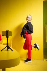 Young fashion girl blogger dressed in red trousers and black jacket takes a selfie on the smartphone standing on a stand on the table in the room with yellow walls and furniture