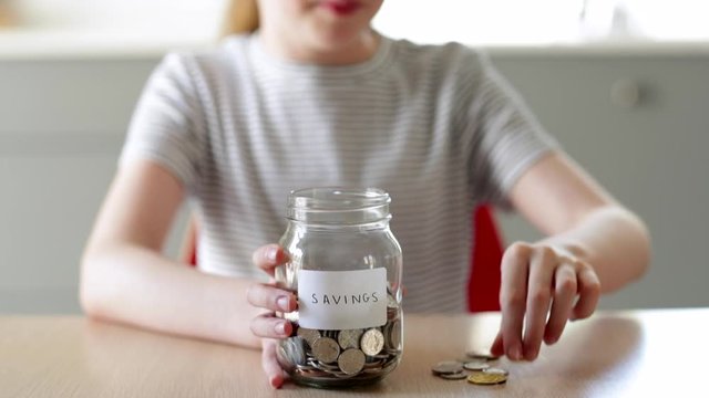 Close Up Of Girl Putting Coins Into Glass Jar Labelled Savings At Home