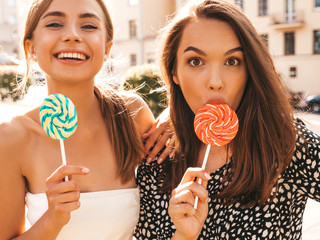 Two young beautiful smiling hipster girls in trendy summer clothes.Sexy carefree women posing on street background.Positive models having fun.Eating candy lollipop