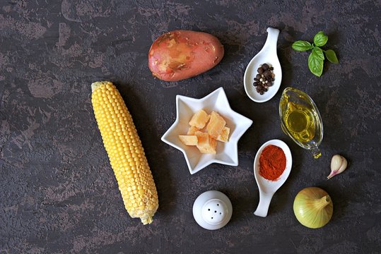 Ingredients for corn soup on a dark background: cob sweet corn, onions, potato, garlic, olive oil, paprika, salt, pepper, green basil, parmesan cheese. Italian food. Top view, copy space.