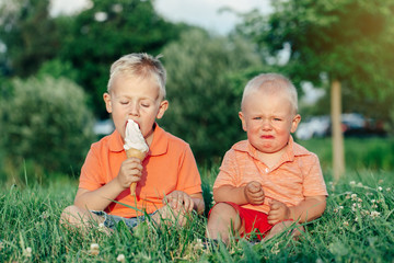 Two Caucasian funny children boys siblings sitting together eating sharing one ice-cream. Toddler younger baby crying and older brother teasing him. Love envy jealous brothers friendship. - 281488091