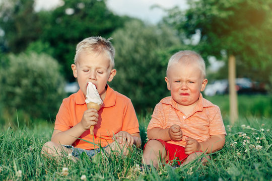 Two Caucasian funny children boys siblings sitting together eating sharing one ice-cream. Toddler younger baby crying and older brother teasing him. Love envy jealous brothers friendship.