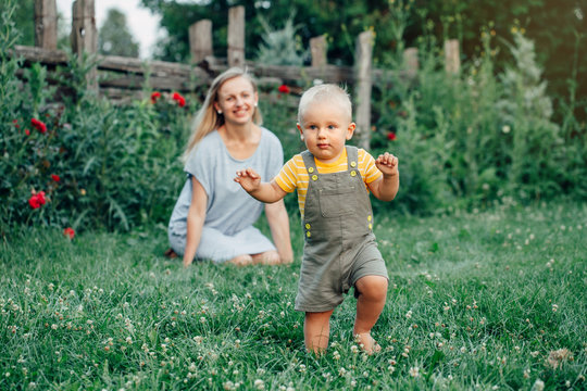 First year child milestone. Baby toddler one year old making his first steps. Caucasian mother watching her son boy walking outside in park on grass on summer day. Happy family lifestyle.