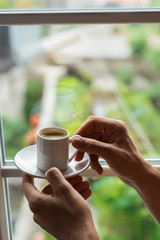 man hands holding small cup of coffee near window in morning sunlight