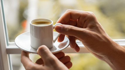 man hands holding small cup of coffee near window in autumn morning