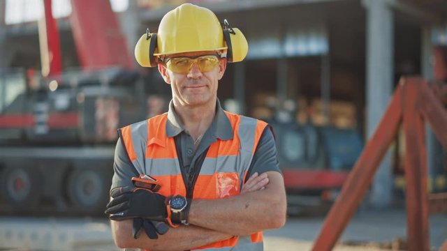 Portrait of Successful Builder / Worker / Contractor Wearing Hard Hat and Safety Vest Standing on a Commercial Building Construction Site, Crosses Arms Confidently. In the Background Crane Machinery