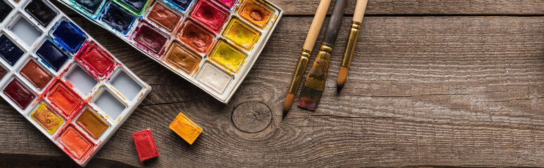 top view of watercolor paint palettes on wooden brown surface with paintbrushes, panoramic shot