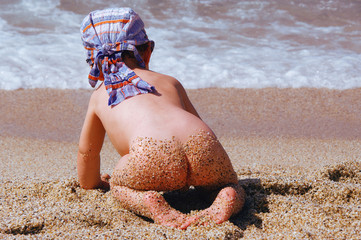 A little boy crawls naked on his knees on the beach and looks at the waves on the sea in the summer...