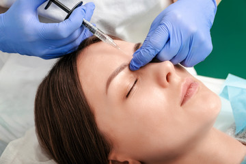 Beautician makes injections between the eyebrows on the face of a beautiful woman. Correction of mimic wrinkles. Cosmetology. Cosmetic procedures in the medical center.