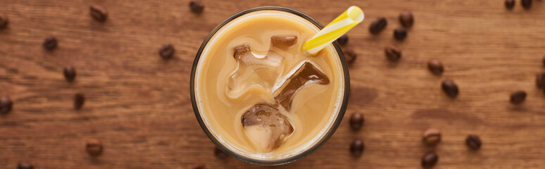 selective focus of ice coffee with straw in glass and coffee grains on wooden table, panoramic shot