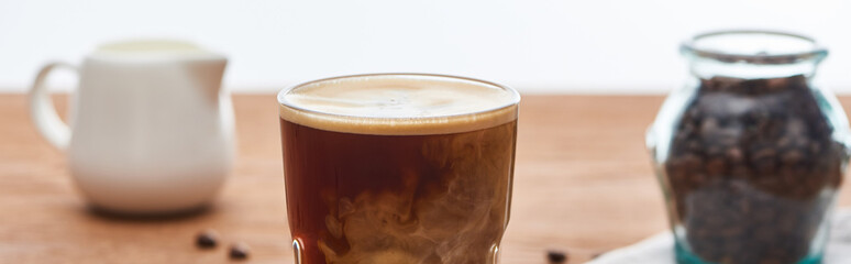 selective focus of ice coffee with milk in glass near milk jug and coffee grains on wooden table isolated on white, panoramic shot