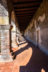 Weathered and worn brick with sandstone springer and keystones in arches form the famous corridors, casting large shadows on red floor tiles at the Mission at San Juan Capistrano, California.