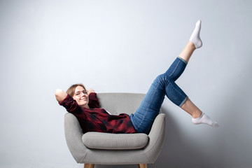 young girl is sitting on a soft comfortable chair against a white wall with her legs raised up, a...