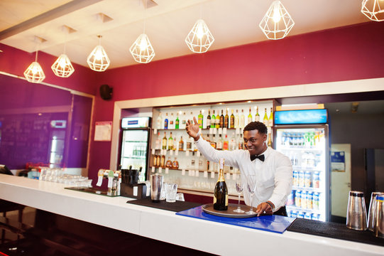 African american bartender at bar holding champagne with glasses on tray.