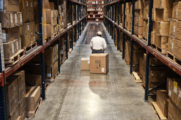 Worker sitting on box between racks of cardboard boxes in distribution warehouse