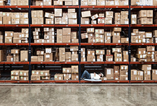 Worker sleeping between cardboard boxes on rack in distribution warehouse