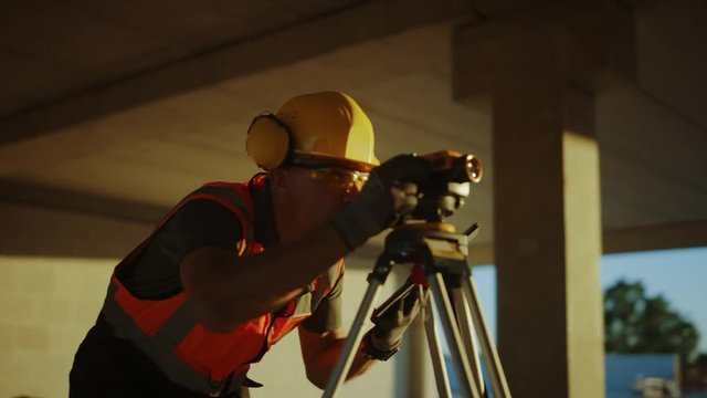 Inside of Commercial / Industrial Building Construction Site: Engineer Surveyor Takes Measures with Theodolite, Using Digital Tablet Computer