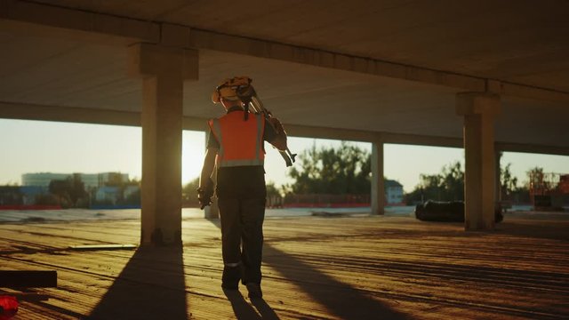 Inside of the Commercial / Industrial Building Construction Site: Engineer Surveyor Finishes Day's Job Carries Tripod Theodolite and Walks into Sunset. In Background Skyscraper Formwork Frames, Crane 