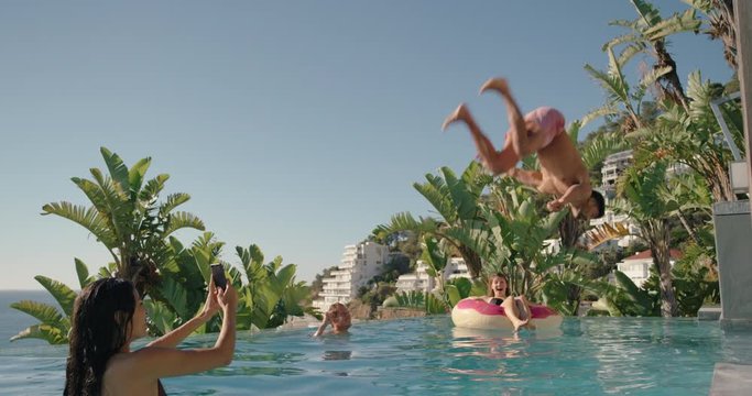 young man doing backflip in swimming pool with woman using smartphone taking video sharing summer vacation with friends on social media having fun enjoying summertime 