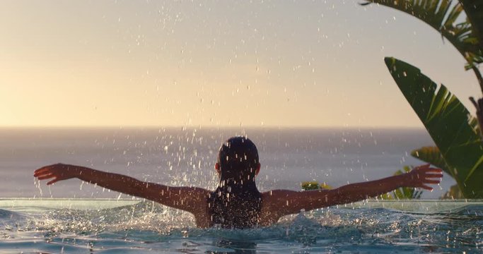 happy woman jumping out of swimming pool water with arms raised enjoying summer vacation in mediterranean hotel resort with beautiful view of ocean at sunset travel freedom 4k