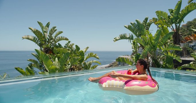 beautiful woman relaxing on pink swim tube floating in swimming pool drinking juice getting sun tan on warm sunny day enjoying summer vacation at luxury hotel resort with view of ocean palm trees 4k