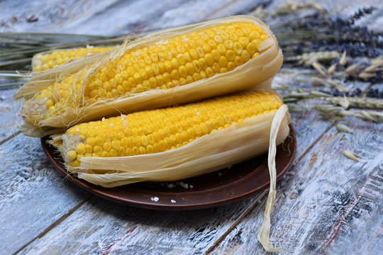 An image of a brown clay plate with three corncobs with salt, a bouquet of ears of corn and lavender on a light multi-colored table
