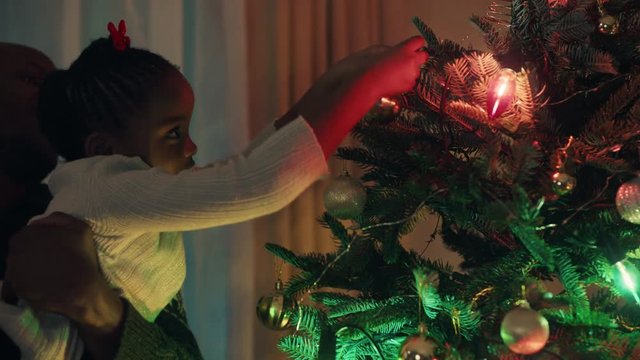 little african american girl with father decorating christmas tree with beautiful ornaments and baubles child helping dad hang festive decorations at home on calm evening 4k footage