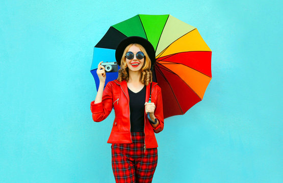Happy smiling woman holding colorful umbrella, retro camera taking picture in red jacket, black hat on blue wall background
