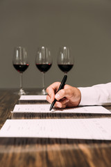 cropped view of sommelier writing in documents at table with wine glasses