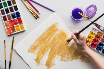 cropped view of artist drawing yellow watercolor brushstrokes on white paper on marble white surface