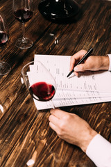 cropped view of sommelier writing in documents at table and holding wine glass