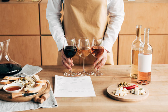 Cropped view of sommelier in apron holding wine glasses near wooden table