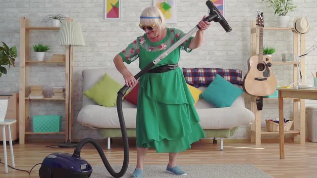 Cheerful happy old woman pensioner with gray hair in glasses playing a vacuum cleaner like a guitar slow mo