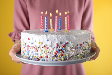 Woman holding birthday cake with burning candles on yellow background, closeup