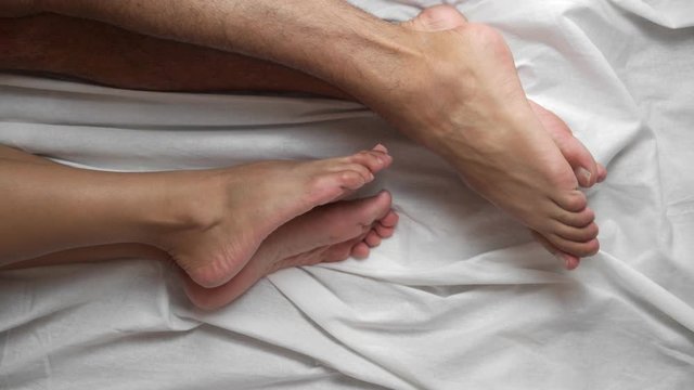 Top view. Female and male legs in bed on a white sheet caress each other. Romance at home. Close-up.