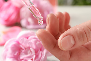 Woman dripping rose essential oil on finger against blurred flowers, closeup