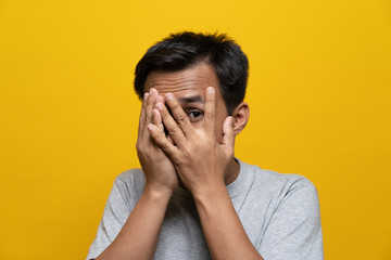 Headshot of scared asian man covering face by his hands and peeking to see with fear.