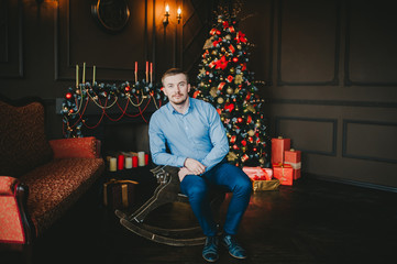 Young man sitting on the wooden horse near Christmas tree with presents and lights in dark living room.