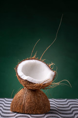 Coconut halves forming a chalice with the outer brown shell and inner white coco on a pattern surface against a dark green background with hairs sticking out. Studio concept of a natural cocktail cup.